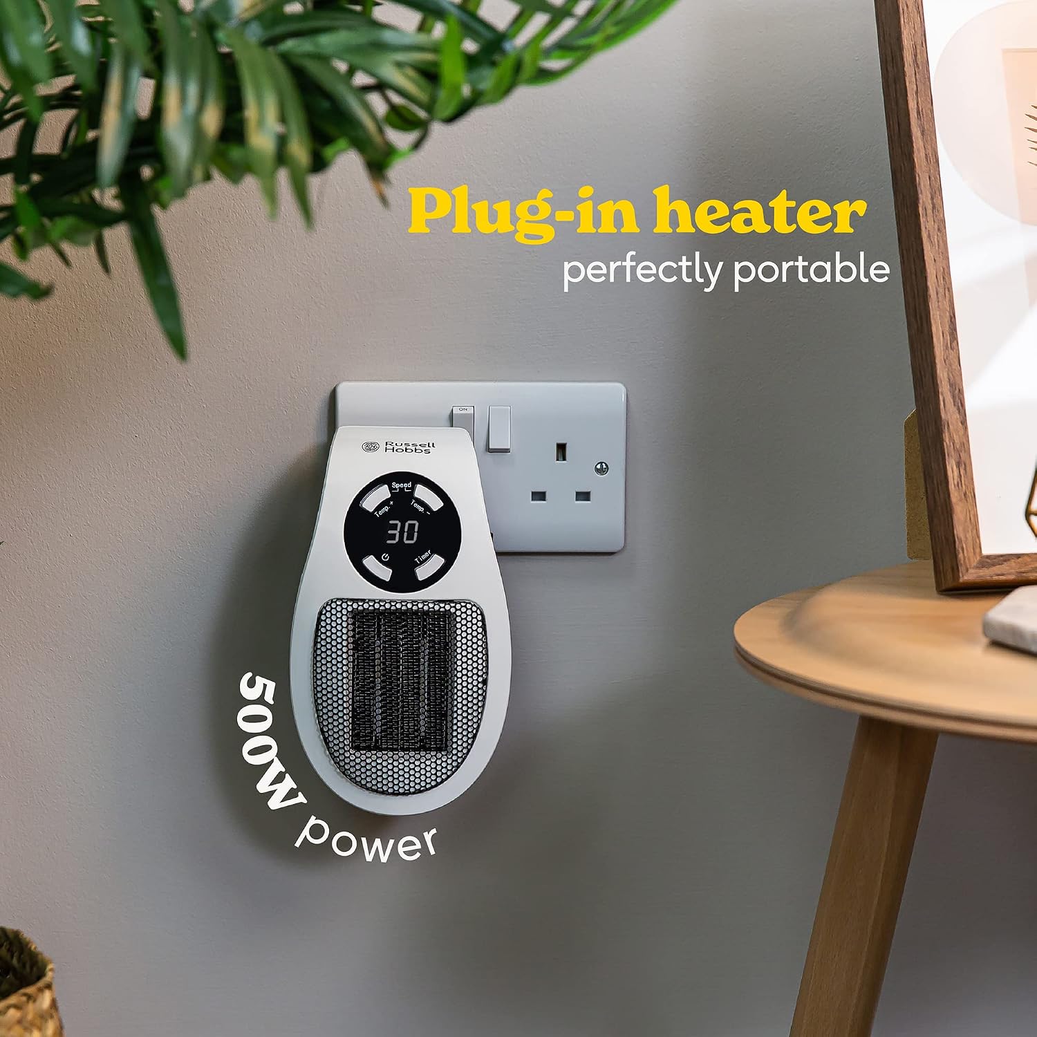 The heater hanging from one outlet of a UK double socket on a living-room wall beside a side table, captioned plug-in heater, perfectly portable, 500W power