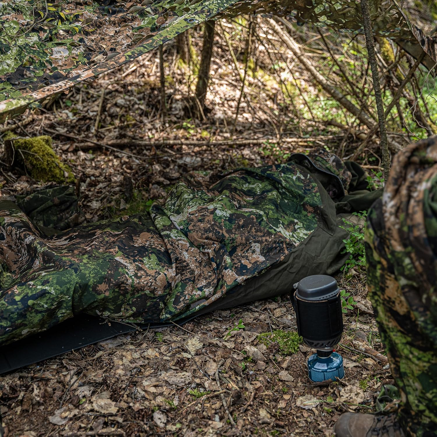Mil-Tec sleeping bag cover laid out on a sleeping mat on the forest floor next to a compact gas stove