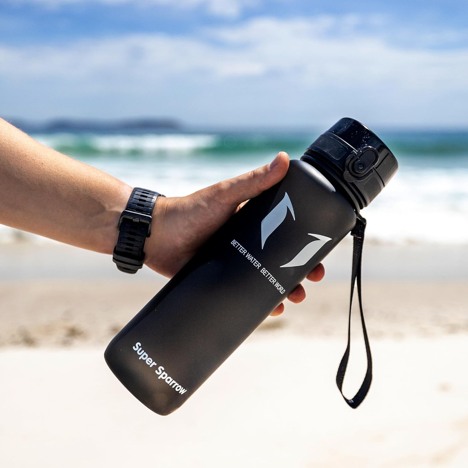 Hand holding a black Super Sparrow bottle by a beach with waves in the background