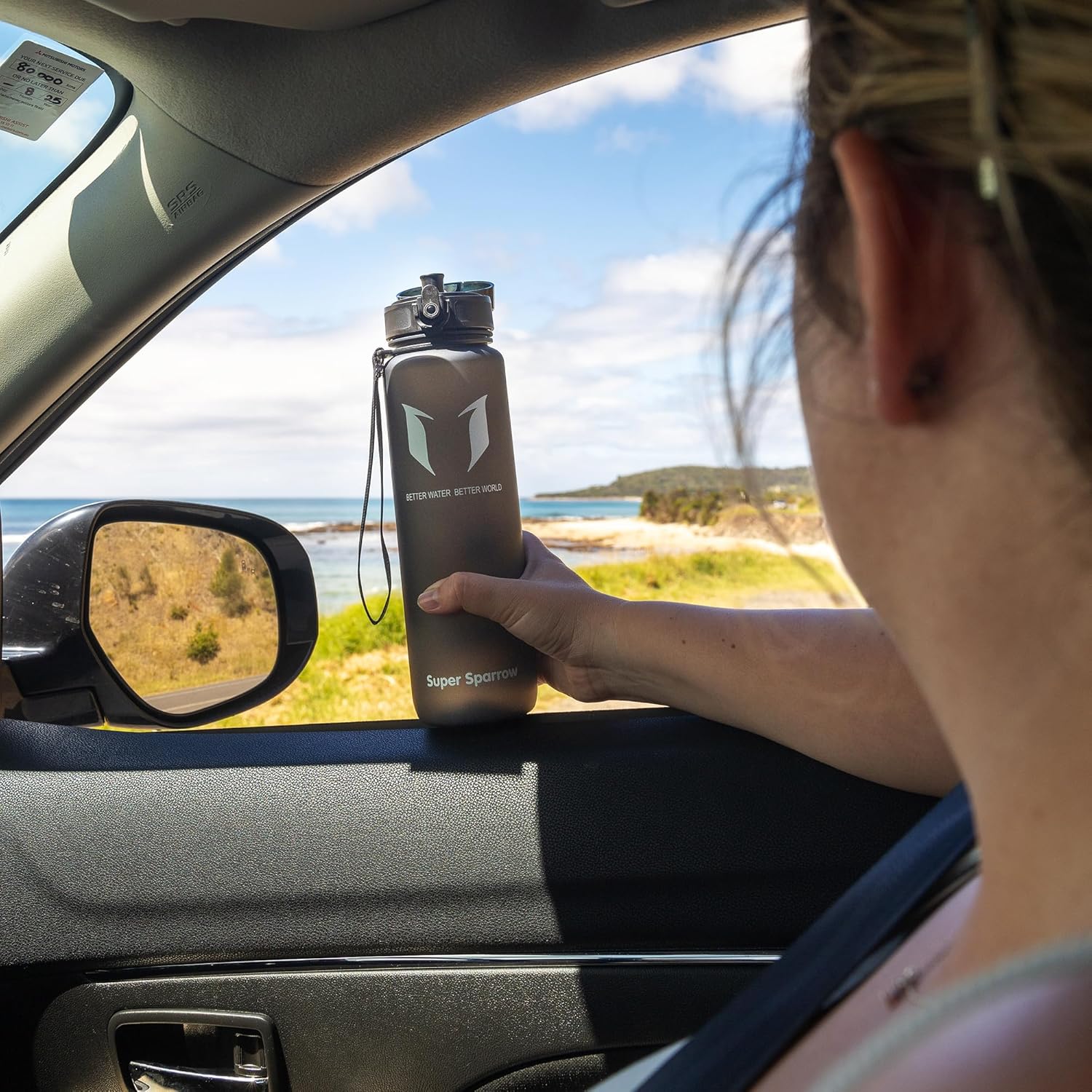 Driver holding a Super Sparrow bottle by the open car window on a coastal road