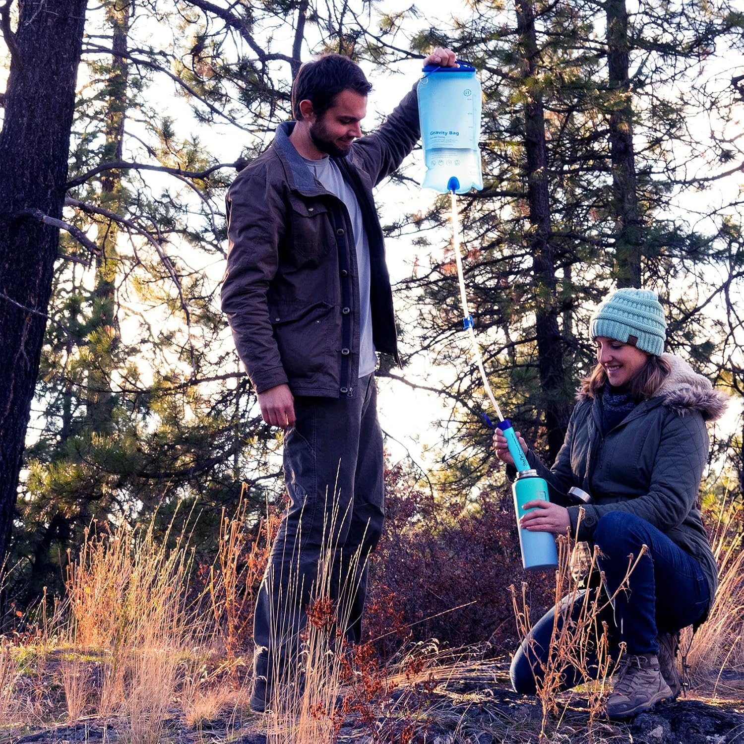 Two campers filling an insulated bottle from a SimPure gravity bag held up by hand in woodland
