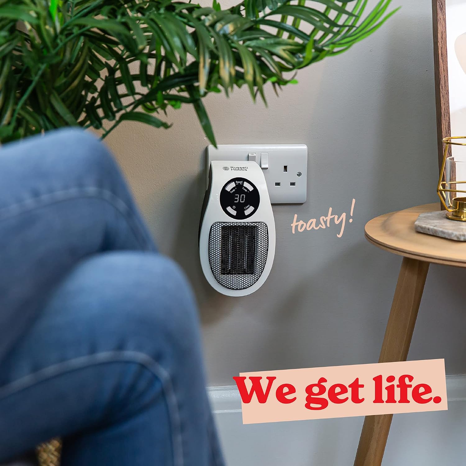 The heater on a living-room wall beside a wooden side table, with a person in jeans in the foreground and the caption toasty