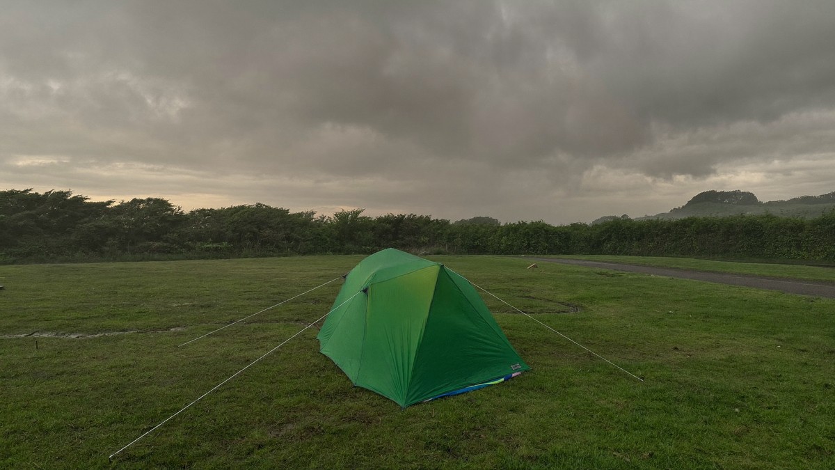 A lone green dome tent pitched on soaked grass at an exposed UK campsite, guylines pulled taut under a heavy grey autumn storm sky