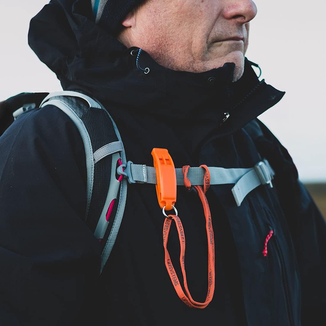 Man outdoors with Lifesystems whistle clipped to backpack shoulder strap at chest height