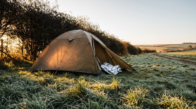 Tent pitched by a hedge windbreak in morning sun with a foil thermal blanket ready for warmth